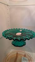 Fenton green glass cake plate with ruffled edges shown on a shelf