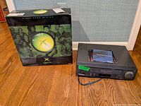 Original Xbox console in its original retail box on hardwood floor next to a Sharp VHS player.