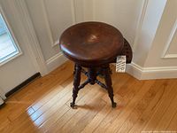 Front angle view of the antique oak swivel piano stool showing the round seat, carved legs, and claw ball feet on hardwood flooring near a door.