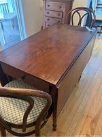 Photo showing antique wooden table with one leaf folded down and two chairs beside it, wood surface and drawer visible.