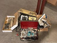 Wide view showing keyboards stacked, coiled cables in a silver case, speakers, a phone handset, and various wired components in cardboard boxes on concrete floor.