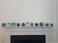 Eleven handcrafted ceramic vases arranged in a row on a white mantle, showcasing various sizes, shapes, and colors including shades of green, blue and gray.