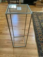 Front and side view of the small narrow square glass table with metal frame showing full height and structure on a hardwood floor next to a patterned rug.