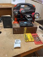 Two cordless drills in front of a black carrying case along with wooden box, yellow and red bit holders, and bits and screws on table.
