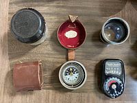 Photo of a compass in leather case, a camera light meter, and camera lenses showing included accessories and tools.