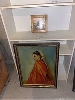 Overall view: large painting leaning below shelf, small framed tile above on shelf