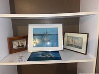 Three framed pictures of boats on a shelf: large one in center, smaller ones to left and right
