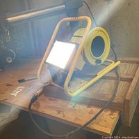 Bright yellow metal framed portable work light turned on, placed on wooden surface, with extension cord attached.