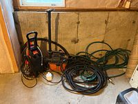 Wide angle image showing the whole lot including pressure washer, shop vac, garden hoses with nozzles, placed on basement floor.