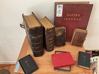 Seven vintage religious books displayed on a wooden table, including two large Holy Bibles, a large burgundy Dante's Inferno, and smaller religious booklets.