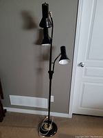 Full view of the black floor lamp standing against a gray wall, showing three adjustable lamp heads and round base with coiled power cord.