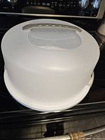 White plastic round cake carrier with dome lid and silver handle seen from above at an angle, sitting on black kitchen stove.