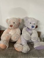 Two teddy bear dolls seated next to each other on a patterned surface, one light pink and the other light purple