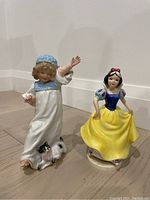 Two porcelain figurines: a child with cat and Snow White, standing on floor against a white wall.