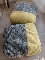Two square knitted fabric floor pouf ottomans, one showing yellow and black/white knit sections on top, on hardwood floor near furniture.