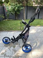 Side view of the black golf push cart with three wheels, showing foldable frame and blue wheels on stone patio next to grass.