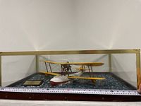 Front angled view of the yellow biplane RCAF Flying Boat model on blue textured water base within glass display case with gold trim, ruler showing approx length under 15 inches.