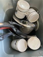 Photo showing four black frying pans/woks stacked with four white ceramic IKEA canisters with locking lids in a sink.