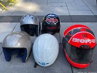 Five helmets displayed on pavement including red 'Beefe' full-face motorcycle helmet, white helmet, silver helmet, black 'Hayashi' and 'Boeri' branded helmet, gray helmet.