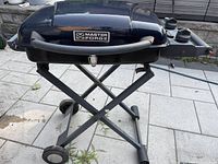 Front view of the black Masterforge portable gas BBQ grill on a foldable metal stand with wheels, placed on an outdoor patio.