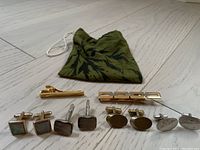 Overview of all cufflinks and tie clips laid out on floor with green cloth pouch behind them