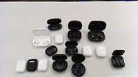 Top-down photo of assorted wireless earbuds in black and white models with charging cases open, showing earbuds inside, placed on a white surface.