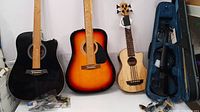 Three instruments side by side: black guitar, sunburst guitar, natural ukulele with case