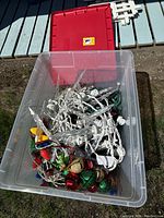 Clear plastic container with white icicle LED light strings and multi-colored bulb string lights with large bulb shapes inside, red storage container lid behind.