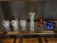 Full view of all six items displayed on glass shelf: three clear floral embossed heart-shaped vases, tall clear swirl vase, small red vase with gold pattern, blue cut glass bowl.
