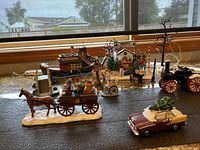 Overall view of village with road holiday-themed figurine set showing horse-drawn cart, vintage car with Christmas tree, steam tractor, Frosty's Christmas Tree building, and other village items on textured surface.