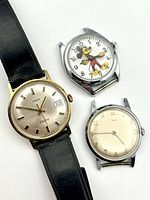 Three men's watches arranged together: Timex Electric with black strap, Timex champagne dial watch, and Mickey Mouse character watch in stainless case without strap.
