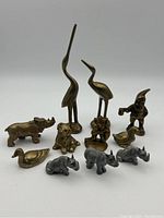 Photo showing 11 brass and pewter figurines including two tall cranes, several animals (rhinoceros, duck, dog, monkey), and a gnome with an axe.
