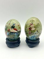 Front view of two natural stone eggs each hand-painted with birds and flowers, displayed on round dark wooden stands.