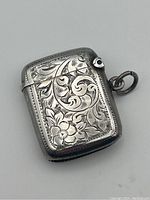 Angled close-up of the front of the intricately engraved sterling silver match vesta showing floral designs and a circular blank cartouche with loop attachment.