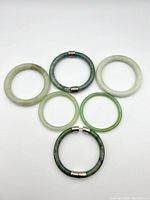 Top down view of the six green stone bangles arranged in a triangle shape on white background. Two bangles have silver clasps, one with the catch broken.