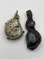 Photo of two pendants: one ammonite fossil in teardrop shape wrapped in silver wire, and one dark blue agate stone wrapped in dark copper and black wire.