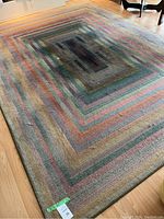 Full view of rectangular multicolor area rug showing concentric rectangular stripe pattern with muted colors including brown, green, blue, red, orange, and purple on hardwood floor.