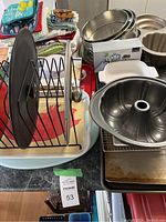 Photo showing stacked metal baking pans including bundt cake pans, wire cooling rack, folded dish towels and cloths piled nearby, Coca-Cola serving tray at bottom, plastic cutting board, and baking molds.