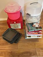 Photo showing pink cupcake storage container, clear cake storage container, 3-tier cupcake stand box, stack of cookbooks, and four square baking pans on floor.
