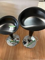Top view of two black bar stools with chrome pedestal bases, showing rounded bucket seats and seat contour details.