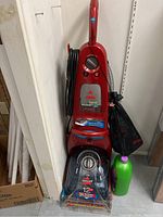 Full view of red Bissell carpet cleaner upright with cord wrapped and detergent bottle beside it