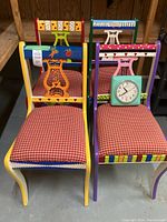 Four colorful hand painted wooden chairs with red and white checkered upholstery and a green wall clock placed on one of the seats.