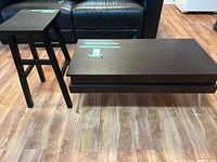 Photo showing coffee table and end table side by side on wood floor in front of a black leather couch.