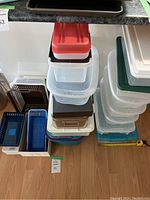Two stacks of assorted plastic and metal containers with lids shown on floor beside cabinet, various sizes and colors including clear, blue, green, and red.