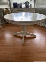 Full view of the round white pedestal dining table with built-in leaf visible in the middle and central pedestal base.
