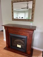 View of electric fireplace in wooden mantel with black insert and faux logs, below an ornate gold framed wall mirror.