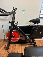 Side view of black Sunny spin bike with red and silver flywheel. Bike placed on wood floor near wall outlet.