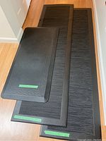 Three black cushioned floor mats stacked on a wooden floor; two longer mats and one smaller mat visible with green measurement labels.