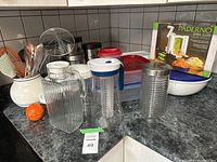 Kitchen countertop with white ceramic utensil holder containing orange and metal utensils, two pitchers (glass and plastic), and boxed Paderno spiral slicer in background