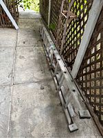 Photo of a large extendable aluminum ladder folded and resting on the ground against a lattice fence outdoors.